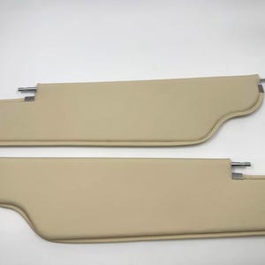 Könnte beinhalten: Zwei beige Autosonnenblenden. Jede Sonnenblende hat eine gebogene Form mit einer Metallstange und Halterung zur Montage. Die Sonnenblenden bestehen aus einem glatten, festen Material und sind so konzipiert, dass sie Sonnenlicht blockieren.