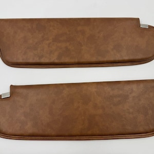 May include: Two brown car sun visors with a textured leather-like appearance. Each visor has a rounded end and a rectangular metal clip. The visors are designed to block sunlight and enhance the interior of a vehicle.