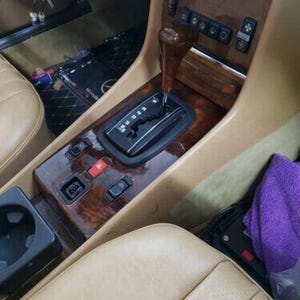 May include: Close-up of a car's interior with a wooden dashboard and a gear shifter. The shifter has a wooden knob and a black plastic gear selector. The dashboard has a variety of buttons and controls.