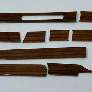 Puede incluir: Un conjunto de 14 piezas de moldura de tablero de instrumentos de imitación de madera para un automóvil. Las piezas son de varias formas y tamaños, y todas están hechas de madera marrón oscura.