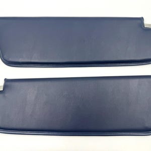 May include: Two navy blue car sun visors with rounded edges and silver metal accents. The visors are made of a smooth, leather-like material. The visors are designed to block sunlight and reduce glare while driving.