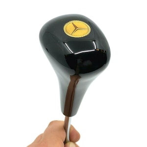 May include: A black gear shift knob with a gold emblem featuring a three-pointed star within a circular border. The knob has a glossy finish and a brown and silver shaft. Automotive accessory.