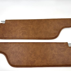 May include: Two brown car sun visors. The visors are rectangular with a curved edge and a metal rod for mounting. The material has a textured appearance. The visors are a warm brown colour.