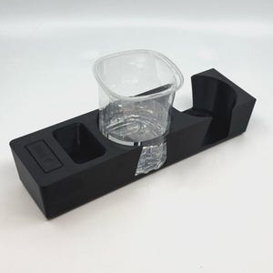 May include: A black plastic cup holder with a clear plastic cup inside. The cup holder has a small compartment on the side.