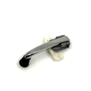 Puede incluir: Una manivela de ventana cromada con un pomo negro y una base de plástico blanco. La manija está en ángulo y parece ser una pieza de repuesto.