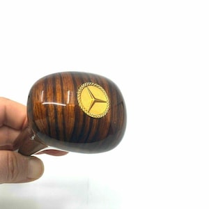 May include: A wooden gear shift knob with a polished finish. The knob features a gold emblem with a three-pointed star design. The wood grain is visible, and the knob is held by a hand.