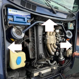 Op de afbeelding: Een open motorkap van een auto met een BMW-motor. Zichtbare onderdelen zijn een blauwe Varta-accu, een gele vloeistofreservoir met een blauwe dop en het motorblok met het BMW-logo. De motorruimte is donkerblauw.