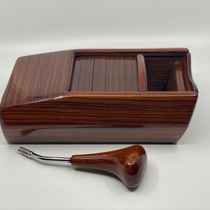 May include: A wooden console with a glossy finish, in a rich, reddish-brown hue and a prominent wood grain pattern. It has a rectangular shape with a compartment and a curved handle with a chrome stem.
