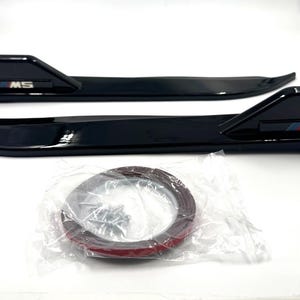 May include: Two glossy black automotive side fender vent trim pieces featuring a raised fin design and the M5 logo with blue, purple, and red stripes. A small plastic bag containing a roll of adhesive tape and several metal screws is included.