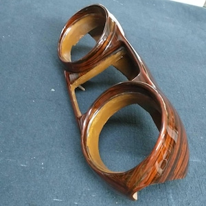 Puede incluir: Un embellecedor de salpicadero de coche de madera con dos aberturas circulares. El embellecedor tiene un acabado brillante con un patrón de vetas de madera en tonos marrón y negro. El interior de las aberturas es de color beige claro.