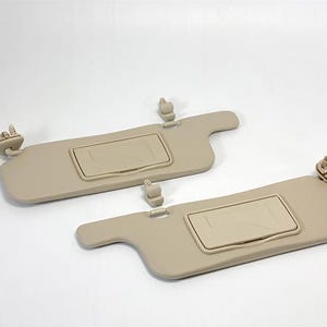 Può includere: Due parasole per auto beige con un vano portaoggetti integrato. I parasole hanno una cornice in plastica beige e un rivestimento in tessuto beige.