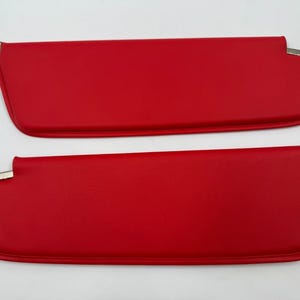 May include: Two bright red car sun visors with chrome-colored metal accents. The visors are rectangular with rounded edges and are made of a smooth, solid material. The visors are designed to block sunlight and improve visibility while driving.