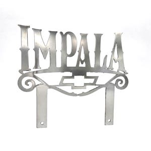 May include: A silver-coloured metal sign featuring the word IMPALA in a stylised font. Below the text is a Chevrolet bowtie emblem flanked by decorative scrollwork. The sign includes two vertical mounting tabs at the bottom with circular holes.