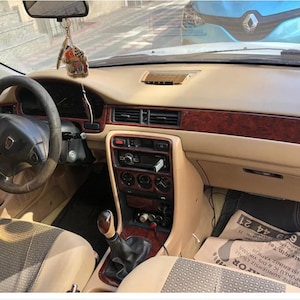 Op de afbeelding: Auto-interieur met beige dashboard, zwart stuurwiel en houtnerf afwerking. Het dashboard bevat een radio, ventilatieopeningen en een versnellingspook. Een papier met tekst is zichtbaar op de vloer.