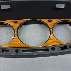 May include: A black plastic dashboard panel with three circular gauge openings. The panel has a woodgrain finish and is designed for a car.