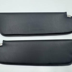 May include: Two black car sun visors with a smooth, leather-like texture. Each visor has a rounded end and a silver-colored metal clip. The visors are designed to block sunlight and enhance driving comfort.