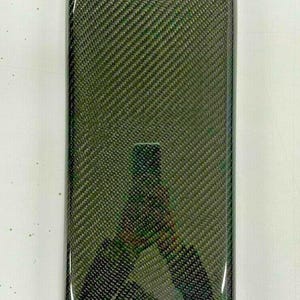 Op de afbeelding: Een zwart carbon fiber middenconsole deksel voor een auto. Het deksel heeft een getextureerd oppervlak en een klein handvat.