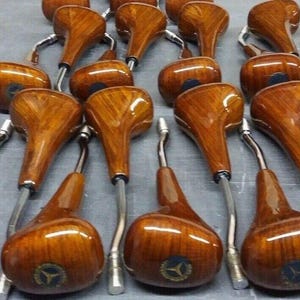 Puede incluir: Primer plano de un grupo de pomos de cambio de marchas de madera con un vástago de metal plateado. Los pomos son de un color marrón intenso con un patrón de veta de madera. Cada pomo tiene un pequeño logotipo en la parte superior.