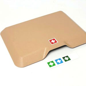 Puede incluir: Un botiquín de primeros auxilios beige con un logotipo de cruz roja. El botiquín tiene una pequeña abertura en la parte delantera. Hay tres pegatinas más pequeñas con cruces blancas sobre un fondo verde, azul y verde.