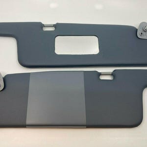 May include: Two grey car sun visors. The top visor has a small rectangular window. The bottom visor has a grey fabric insert.