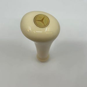 Peut inclure: Pommeau de levier de vitesses ivoire avec un emblème doré représentant une étoile à trois branches dans un cercle. Le pommeau a une finition lisse et brillante et un design incurvé et ergonomique.