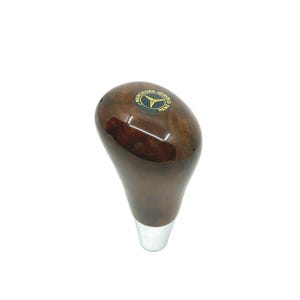 May include: A brown wooden gear shift knob with a silver metal base. The knob features a black and gold Mercedes-Benz logo.