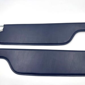 Op de afbeelding: Twee marineblauwe zonnekleppen voor auto's. Elke zonneklep heeft een gebogen vorm met een metalen staaf en beugel voor montage. De zonnekleppen zijn gemaakt van een glad, effen materiaal.