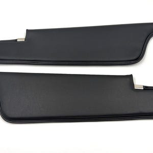 May include: Two black car sun visors. Each visor has a rectangular shape with a curved end and a metal clip. The visors are made of a smooth, solid black material.