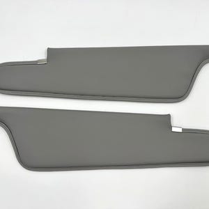 May include: Two gray car sun visors. Each visor has a curved shape with a rectangular main section and a rounded end. The visors are made of a smooth material and have a stitched edge. Metal clips are visible.