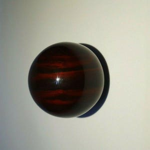 Könnte beinhalten: Runder, dunkelbrauner Türknauf aus Holz mit polierter Oberfläche. Der Knauf hat eine kugelförmige Form und ist auf einer weißen Oberfläche montiert. Die Holzmaserung ist sichtbar und verleiht dem Design Textur.