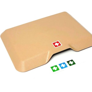 Puede incluir: Un botiquín de primeros auxilios beige con un logotipo de cruz roja. El botiquín tiene una parte superior curva y una pequeña abertura en la parte delantera. Cuatro pegatinas con cruces blancas sobre fondos de colores diferentes se muestran debajo del botiquín.