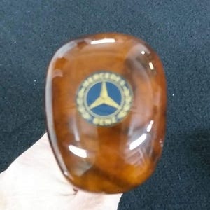 May include: A close-up of a brown wooden gear shift knob with a gold Mercedes-Benz logo. The logo is a three-pointed star inside a circle with the words "Mercedes-Benz" around it.