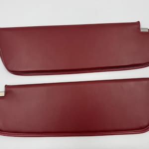 May include: Two burgundy-colored car sun visors. Each visor has a rectangular shape with rounded edges and a silver-colored metal bracket. The visors are made of a smooth, leather-like material.
