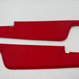 May include: Two bright red car sun visors. Each visor has a rectangular shape with a curved end and a metal clip. The visors are made of a smooth material and are set against a white background.