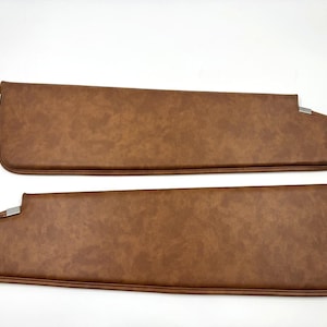 May include: Two brown car sun visors. The visors are rectangular with rounded edges and a textured surface. Each visor has a silver metal bracket and a metal rod for mounting. The visors are made of a leather-like material.