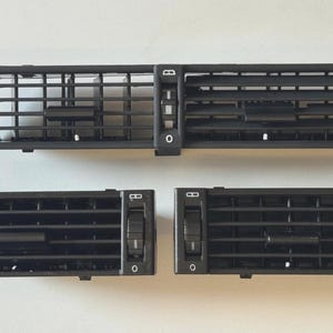 Puede incluir: Un juego de tres rejillas de ventilación negras para coche. Las rejillas tienen forma rectangular con lamas horizontales y verticales. La rejilla central es más larga y tiene un panel de control.