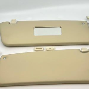 May include: Two beige car sun visors with a rectangular mirror and mounting hardware. The visors are made of a smooth material and have a curved shape. The mounting hardware includes a metal rod and a plastic clip.