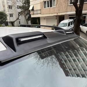 May include: Black car spoiler with an integrated LED light bar. The spoiler is mounted on the roof of a white car. The image is taken outdoors, with buildings and other cars in the background.