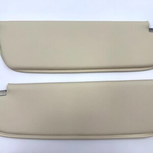 May include: Two beige car sun visors. Each visor has a rounded shape with a metal bracket on one end. The visors are made of a smooth, leather-like material and are designed to block sunlight.