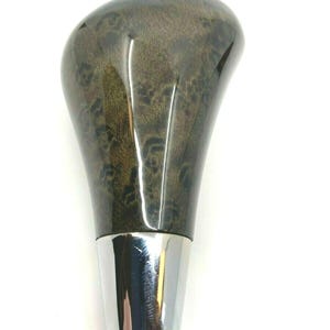 Puede incluir: Un pomo de palanca de cambios de coche con un patrón de veta de madera oscura y una base cromada pulida. El pomo tiene una forma suave y ergonómica y un acabado brillante, que refleja la luz. El diseño combina la estética de la madera natural con un acento metálico moderno.