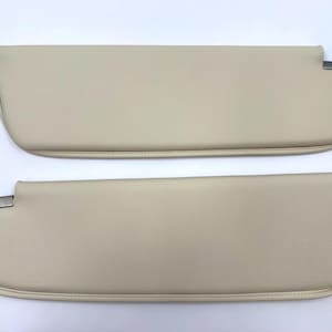 May include: Two beige car sun visors. Each visor has a rectangular shape with rounded edges and a metal bracket. The visors are made of a smooth, leather-like material and are designed to block sunlight.