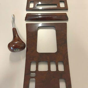 Puede incluir: Un conjunto de piezas de moldura interior de coche de madera marrón con una perilla de cambio de marchas. Las piezas de moldura tienen un patrón de veta de madera y están diseñadas para adaptarse al tablero de instrumentos y la consola central de un coche.