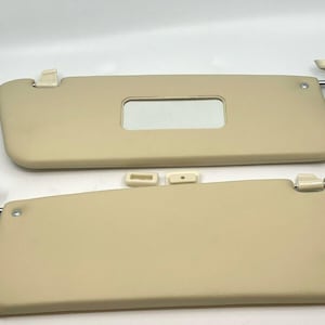 May include: Two beige car sun visors with a small rectangular mirror on the top visor. The visors have a metal arm with a plastic clip.