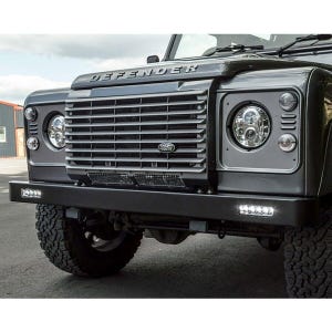 Puede incluir: Un Land Rover Defender gris con un parachoques delantero negro y una parrilla plateada. La parrilla tiene el logotipo de Land Rover en el centro. Los faros están encendidos y el coche está aparcado en una superficie pavimentada.