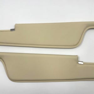 May include: Two beige car sun visors. Each visor has a curved, padded design with a stitched edge and a metal clip for attachment. The visors are a light tan color and appear to be made of a smooth material.