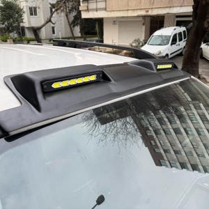 Puede incluir: Accesorio negro para el techo del coche con luces LED integradas. El accesorio es negro y tiene un diseño elegante, con una serie de luces LED amarillas brillantes. Las luces están dispuestas en fila y son visibles sobre el fondo negro. El parabrisas del coche es visible.