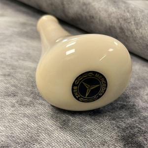 Op de afbeelding: Crèmekleurige, peervormige versnellingspookknop met een glanzende afwerking. De basis heeft een zwart en goud Mercedes-Benz logo. De knop is ontworpen voor de versnellingspook van een voertuig.