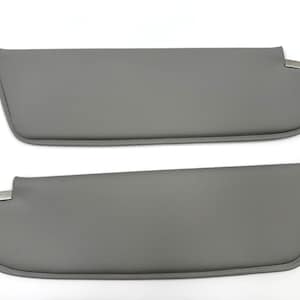 May include: Two gray car sun visors. Each visor has a rounded end and a rectangular metal clip. The visors are made of a smooth, solid gray material and are designed to block sunlight.
