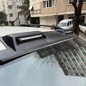 May include: Black car spoiler with an integrated LED light bar. The spoiler is mounted on the roof of a white car. The image is taken outdoors, with buildings and other cars in the background.