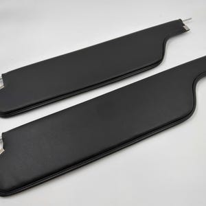 May include: Two black car sun visors. Each visor is rectangular with a curved end and a metal rod for mounting. The visors are made of a smooth, matte material and are positioned on a white background.